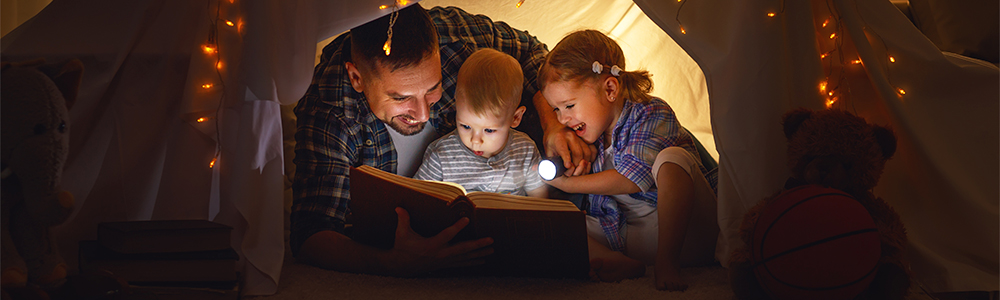 Isä lukee kijraa kahden lapsen kanssa viltin alla.